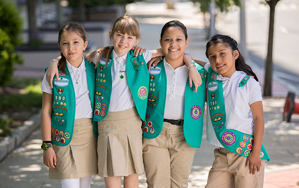 group of junior girl scouts in uniform green vest with patches and badges with arms around each other smiling